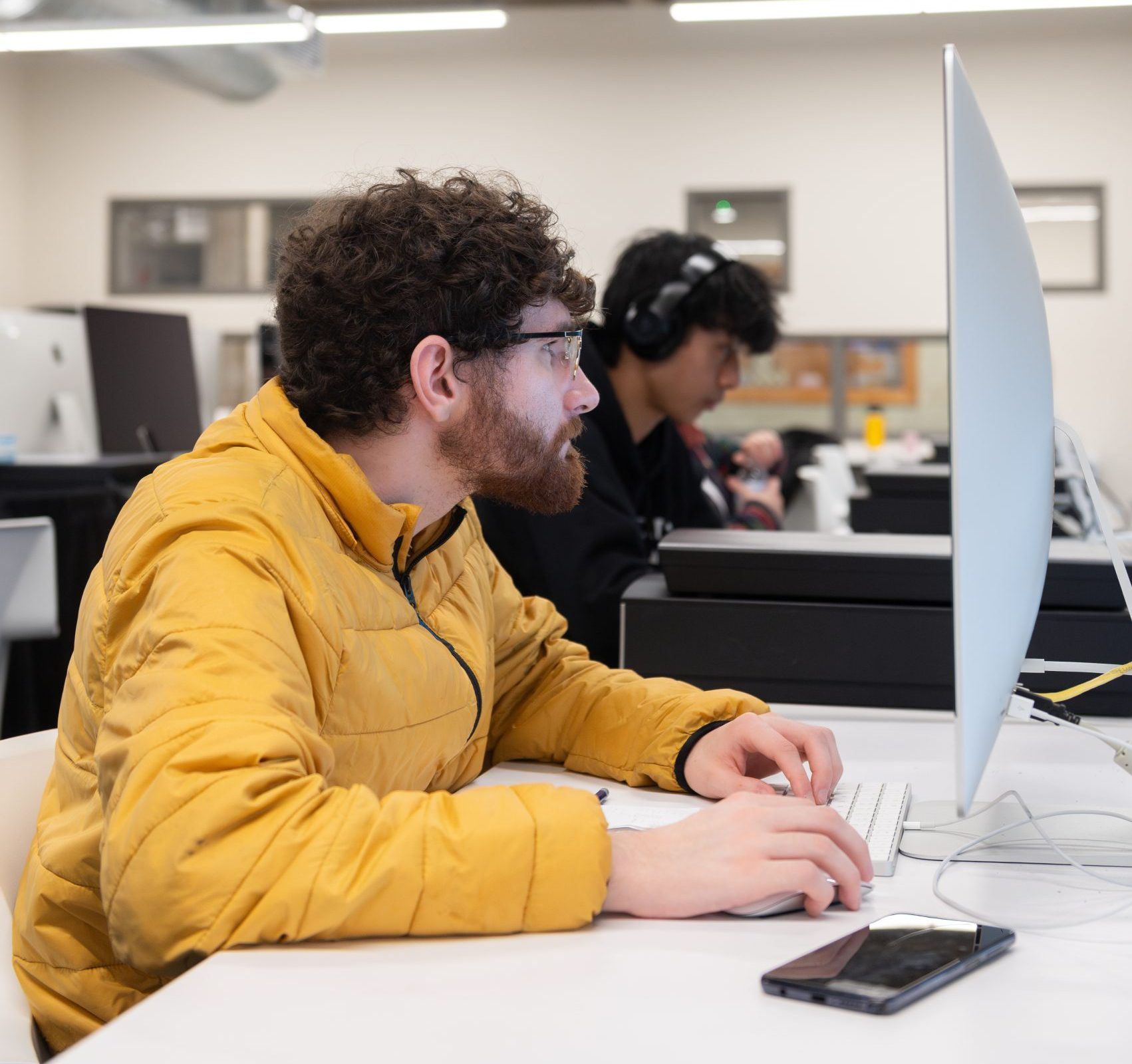 Person wearing a yellow jacket working on an iMac in a modern computer lab, with another person seated and working at a computer in the background.